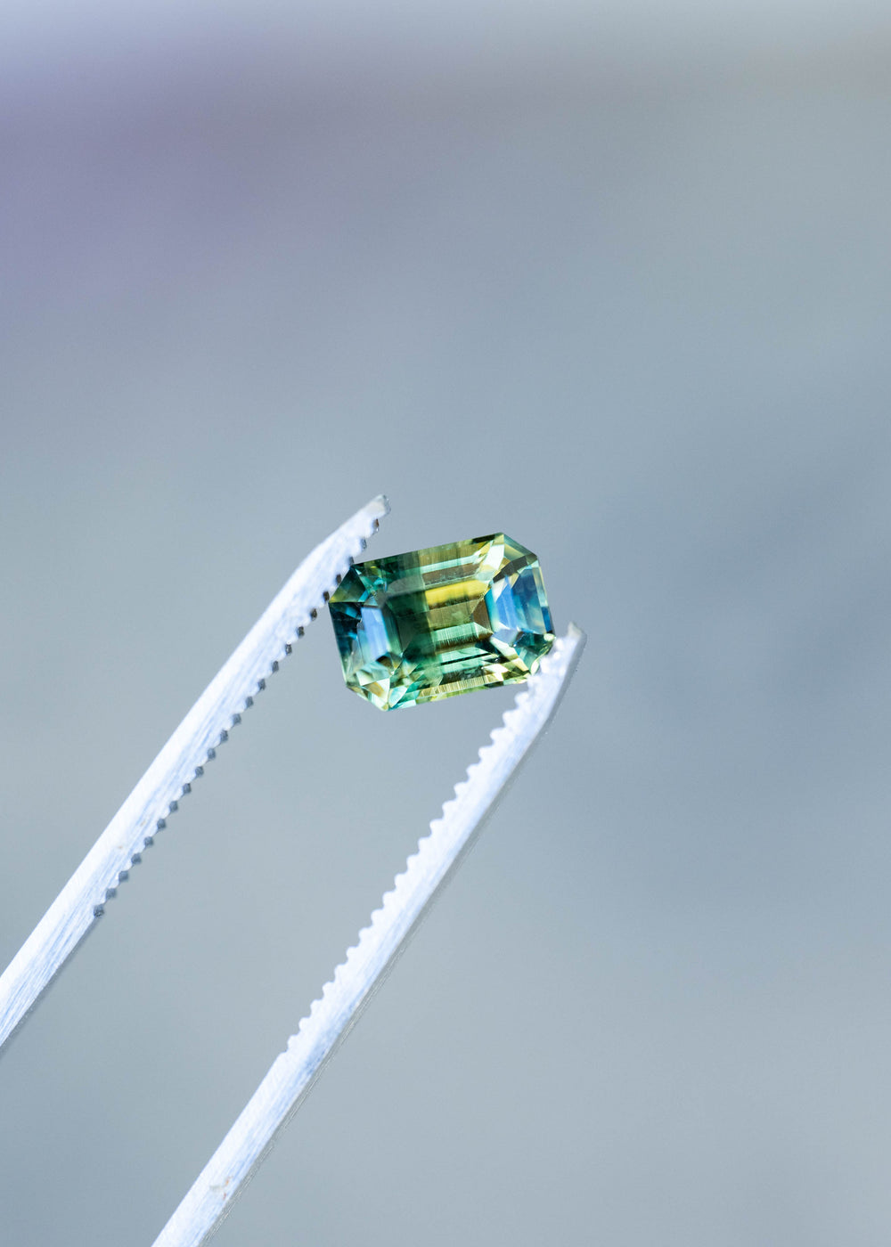Close-up photograph of a 2.16ct untreated Madagascar parti sapphire held in jeweler's tweezers. Precision step facets reveal contrasting bands of yellow, teal green and blue.