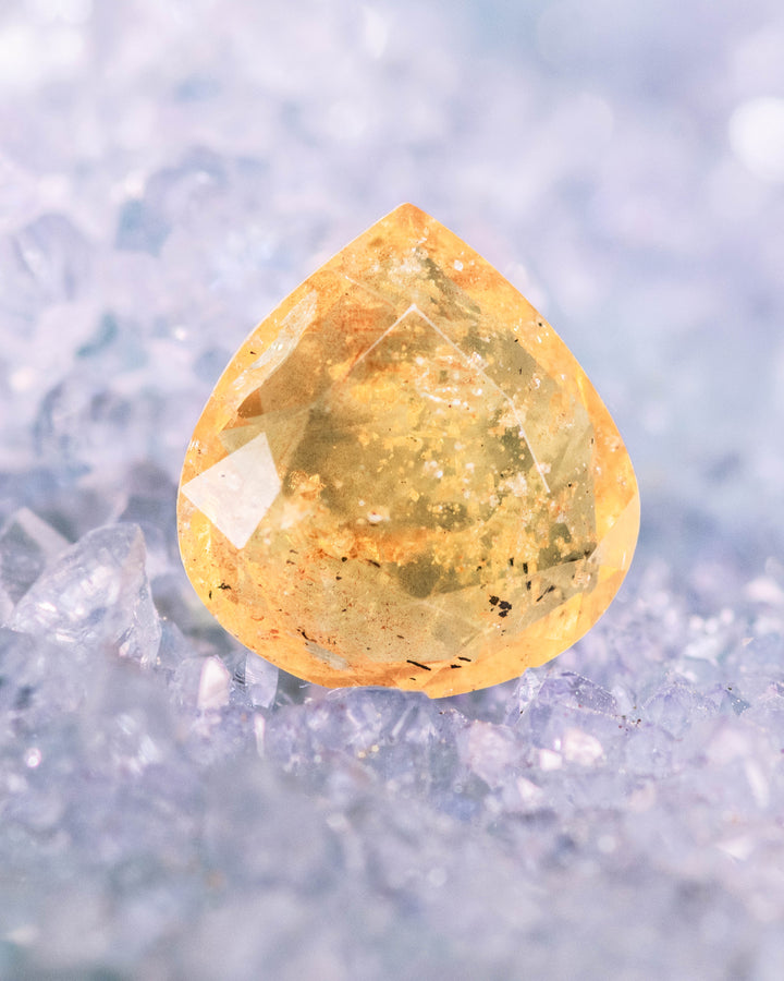 2.17CT yellow brown orange sapphire placed on a bed of icy blue crystals, highlighting its radiant warm glow and internal texture within a custom wide pear cut, unheated sapphire from Umba River, Tanzania.