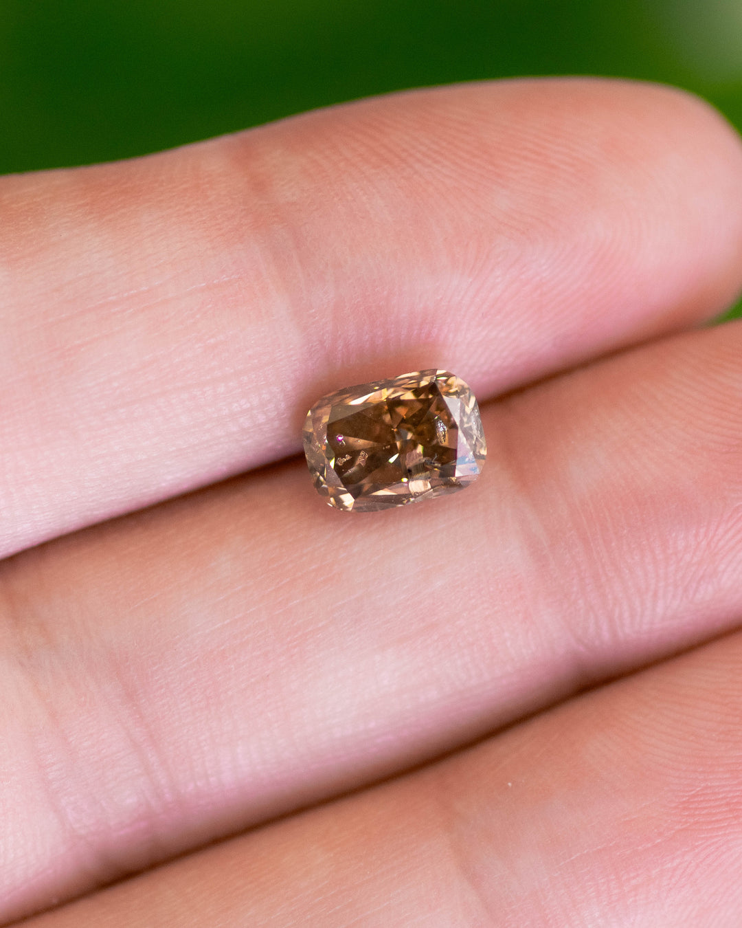 Macro shot of a 2.20 carat fancy brown diamond from Argyle Mines, Australia, resting on a fingertip. The elongated cushion cut reveals warm chocolate-brown tones, strong blue fluorescence, and I2 clarity under natural light.