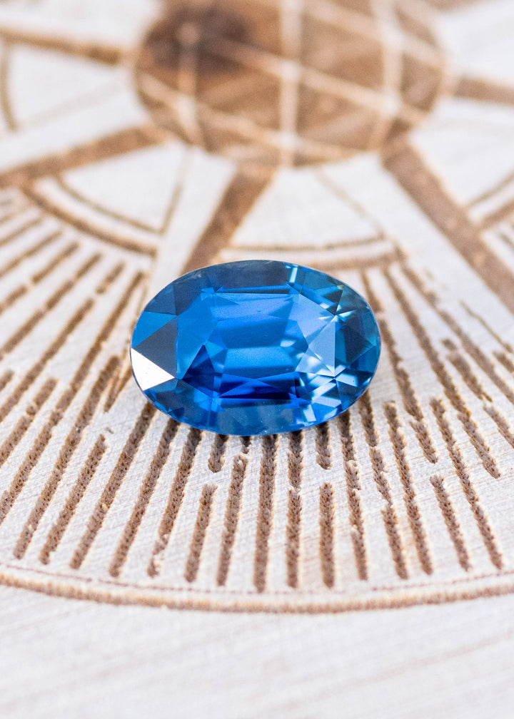 2.29ct Deep oceanic blue oval mix cut sapphire resting on etched wood surface under natural lighting.