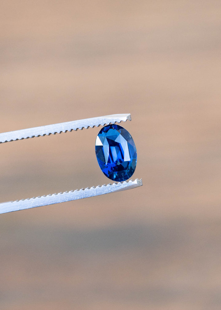 2.29ct Deep oceanic blue oval mix cut sapphire held in gem tweezers under natural lighting.