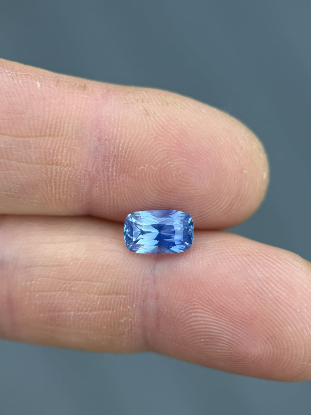 2.30 carat cornflower blue sapphire shown resting on fingertip, displaying precision elongated cushion mixed cut facets and natural brilliance; untreated Madagascar sapphire gemstone.