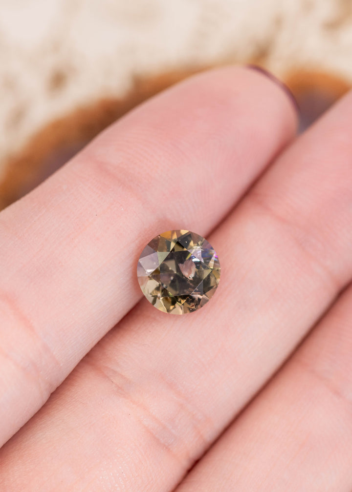2.33 carat Tanzanite resting on fingertip to show scale and brilliance, featuring an antique style round transitional brilliant cut with teal, green, purple, and golden yellow tones