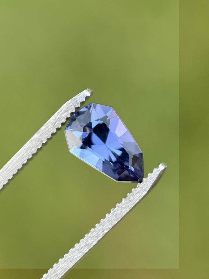 2.42 carat tanzanite gemstone held by precision tweezers outdoors, displaying vivid blue-purple color play and precise shield mix cut facets, ethically sourced from Merelani Hills, Arusha, Tanzania.