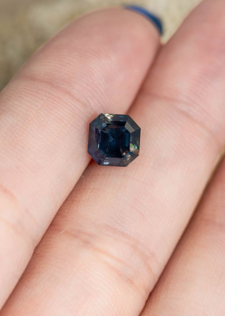 Loose color change sapphire resting on fingertips, showing the Asscher cut step facets and subtle shift from teal blue to purple gray tones.