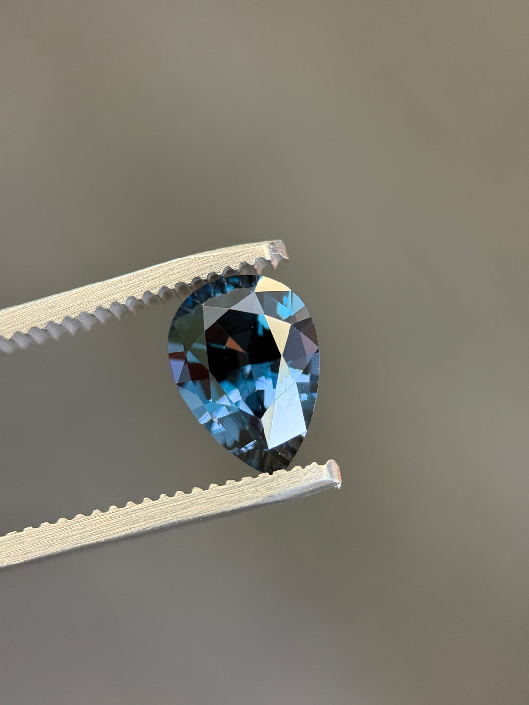A faceted pear-shaped blue gemstone held between white metal tweezers against a plain gray background.