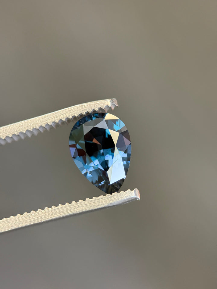 A faceted pear-shaped blue gemstone held between white metal tweezers against a plain gray background.