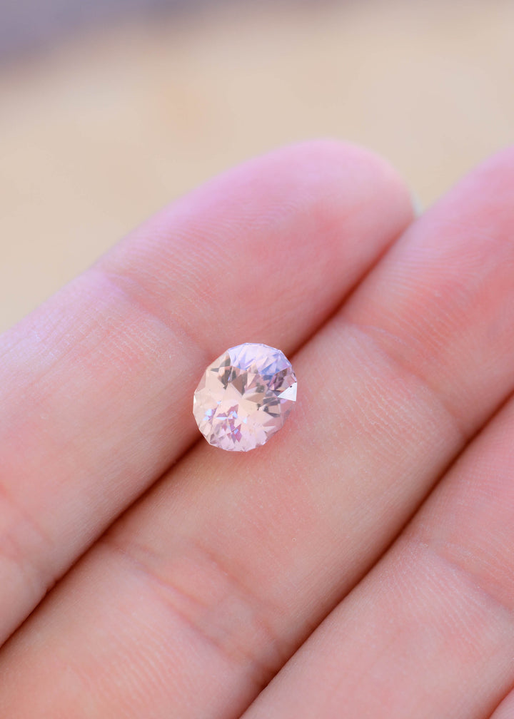2.50 carat pale peach pink sapphire resting on fingertips for scale, showing its elongated geometric oval shape, soft natural color and bright faceting.