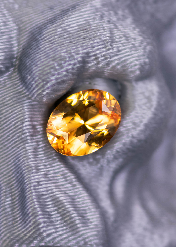 2.53 carat orange yellow sapphire in a mixed brilliant oval cut resting on textured fabric, showing bright golden fire and faceted brilliance from Sri Lanka