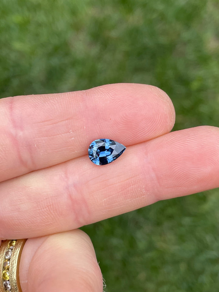 Natural 2.54 carat sapphire in teal, blue, and green hues, pear brilliant cut, ethically sourced from Madagascar, held between fingers outdoors.