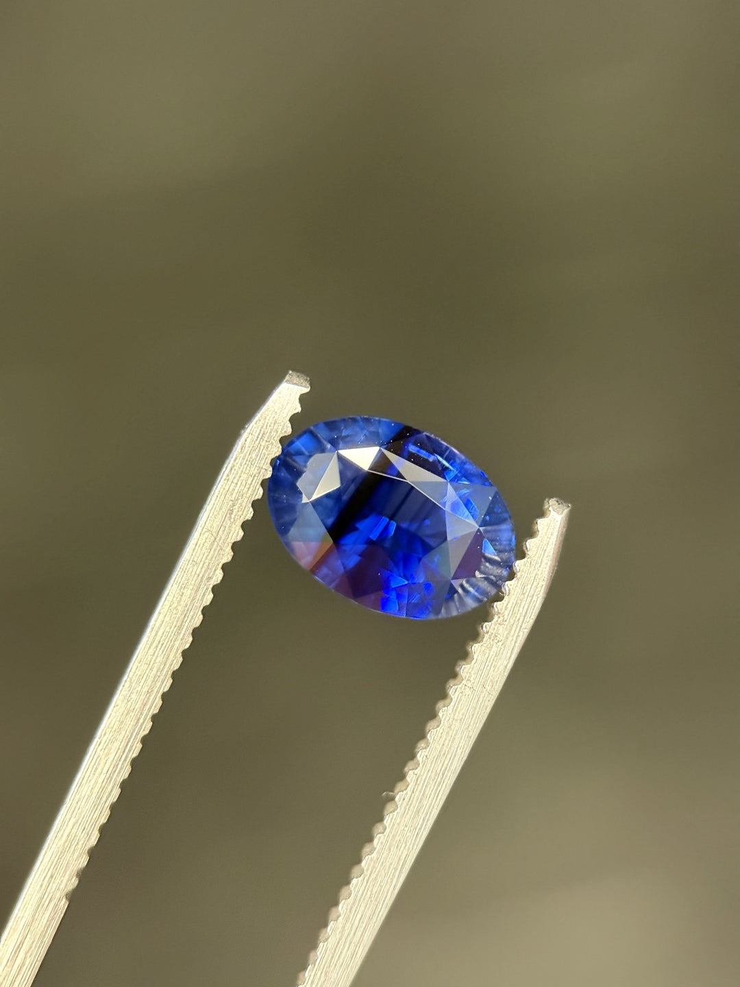 Striated royal blue parti sapphire, 2.56 carats, held in gem tweezers against a neutral background, displaying intense royal blue flashes and precision oval mixed cut brilliance from Diego, Madagascar, untreated.