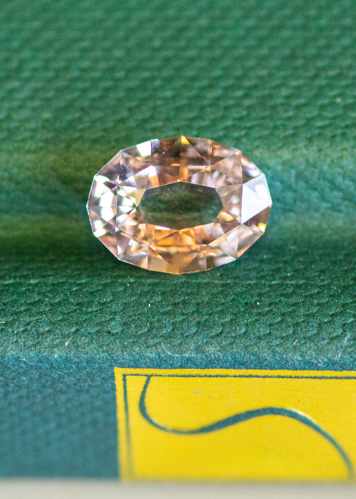 2.56ct blush peach pink antique style oval cut sapphire from Madagascar resting on a textured green book cover.