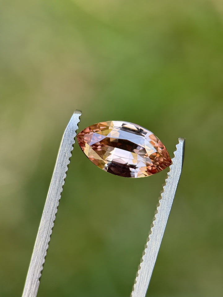 2.57ct marquise-cut sapphire in peach, pink, and rose-champagne tones held in gem tweezers outdoors, highlighting brilliance, facet pattern, and natural color zoning; precision-cut, untreated stone from Madagascar.