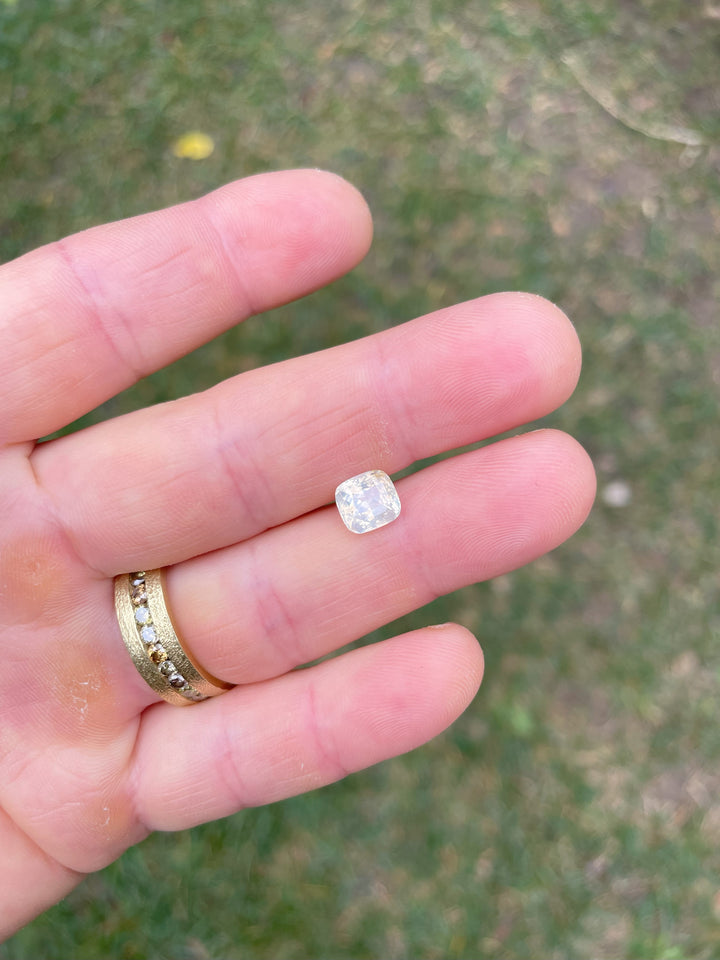 A 2.60-carat opalescent pale yellow sapphire in a custom cushion cut held between fingers, showcasing its unique glow and clarity against a natural outdoor background.