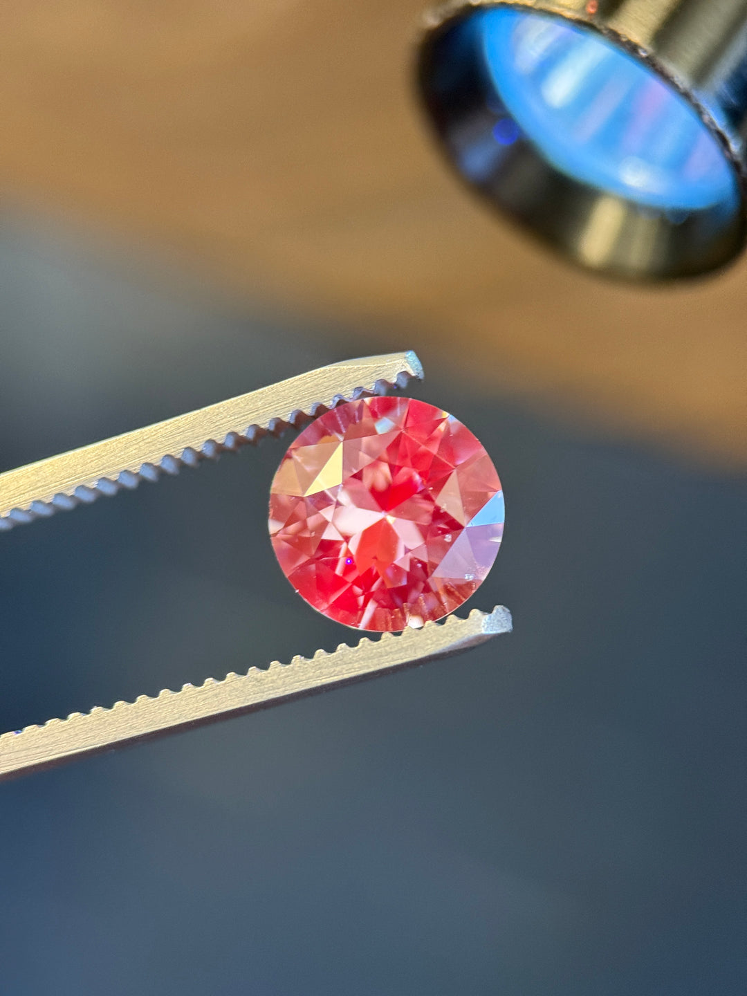 Precision antique transitional round cut sapphire showing a cool steely grey color while held in gemstone tweezers against a blurred green background.