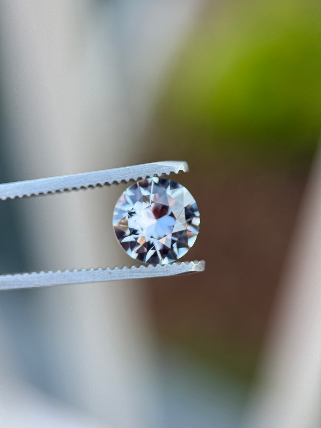 Color-change sapphire under strong light in gemstone tweezers, displaying bright violet and reddish undertones in a 2.60CT antique round transitional cut.