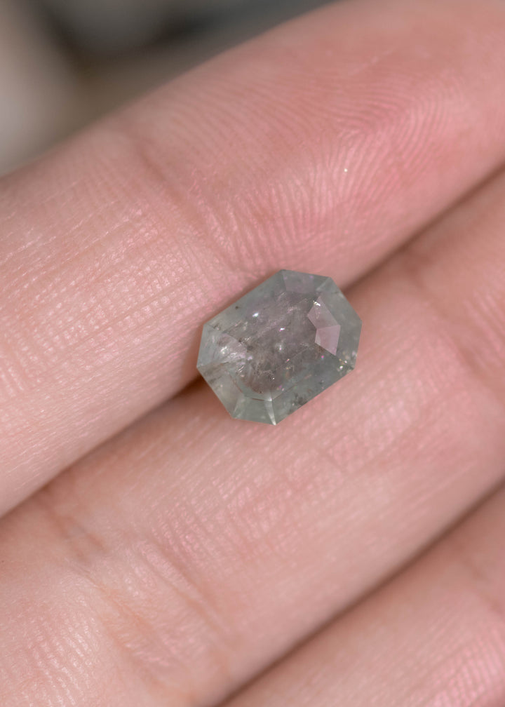 Seafoam green geometric portrait cut sapphire on fingertip for scale, showing pale green body color, internal crystalline inclusions, and a sculptural octagonal outline.