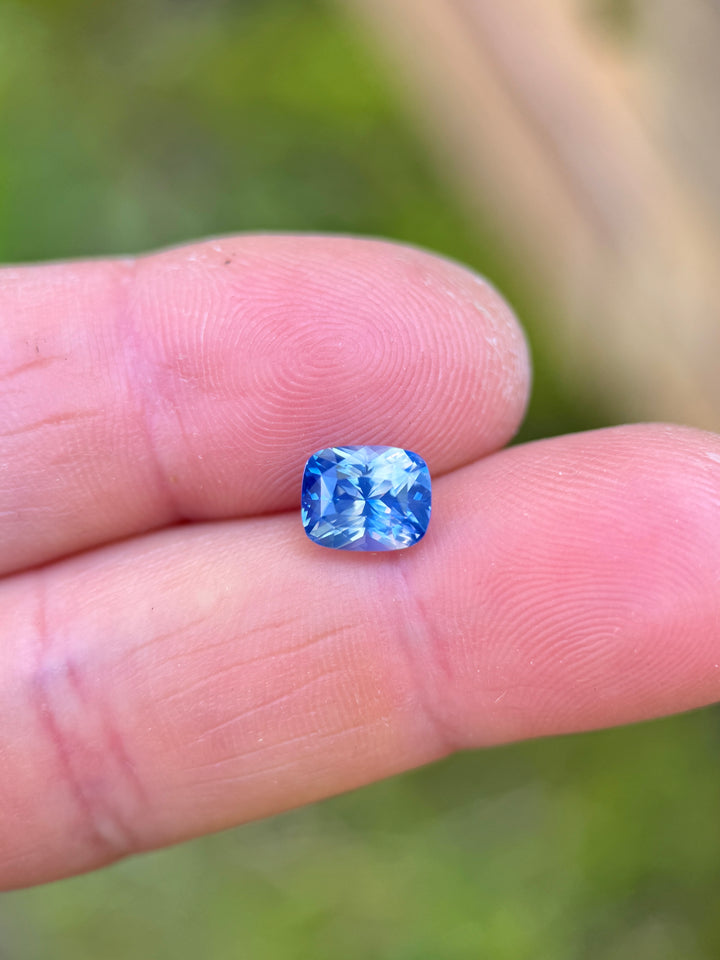 2.64 carat opalescent parti blue sapphire displayed on fingertips, revealing soft pastel blue and white reflections and the precise craftsmanship of the mix cushion cut, untreated sapphire from Sakaraha, Madagascar.