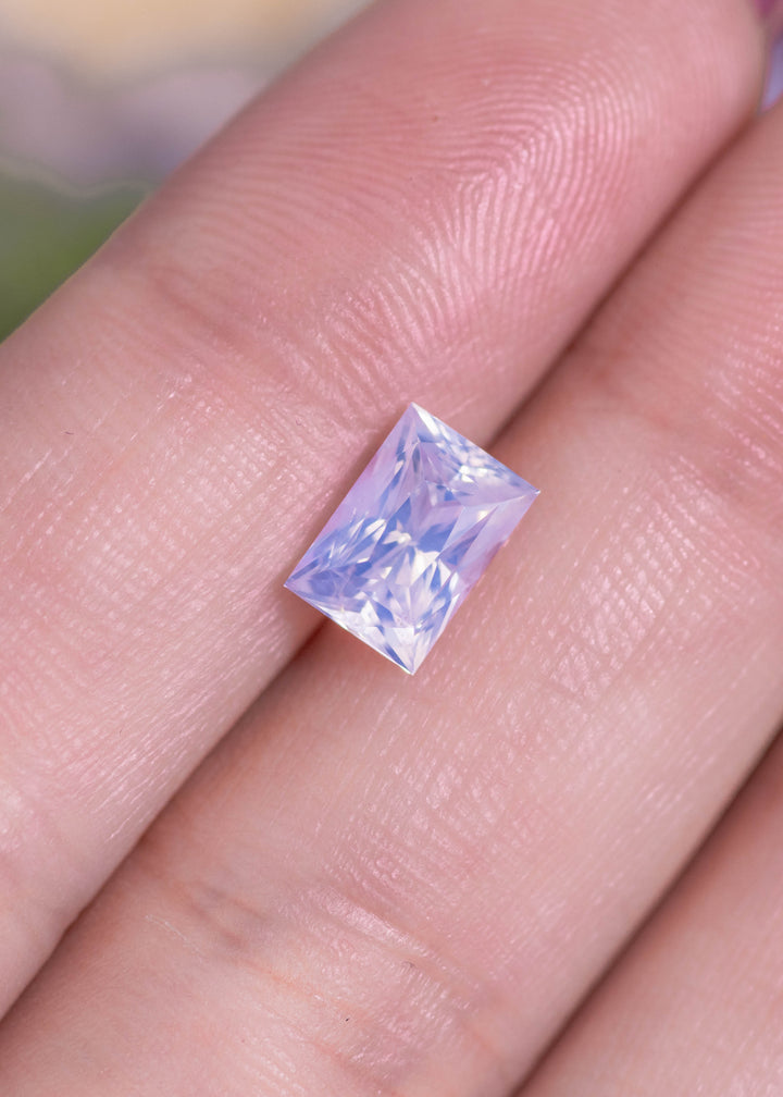 Loose opalescent white sapphire resting on a fingertip for scale, showing the elongated princess cut pattern and a subtle lilac glow in the light.