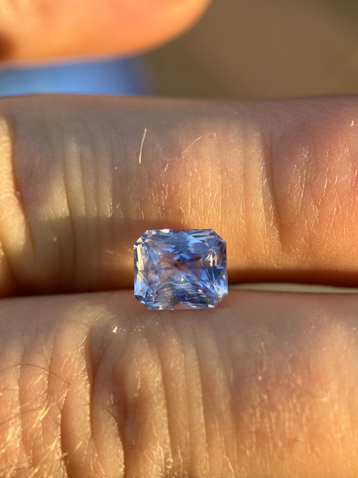 Radiant cut sapphire in lavender, violet, and blue hues weighing 2.70 carats, displayed on fingers in natural sunlight.
