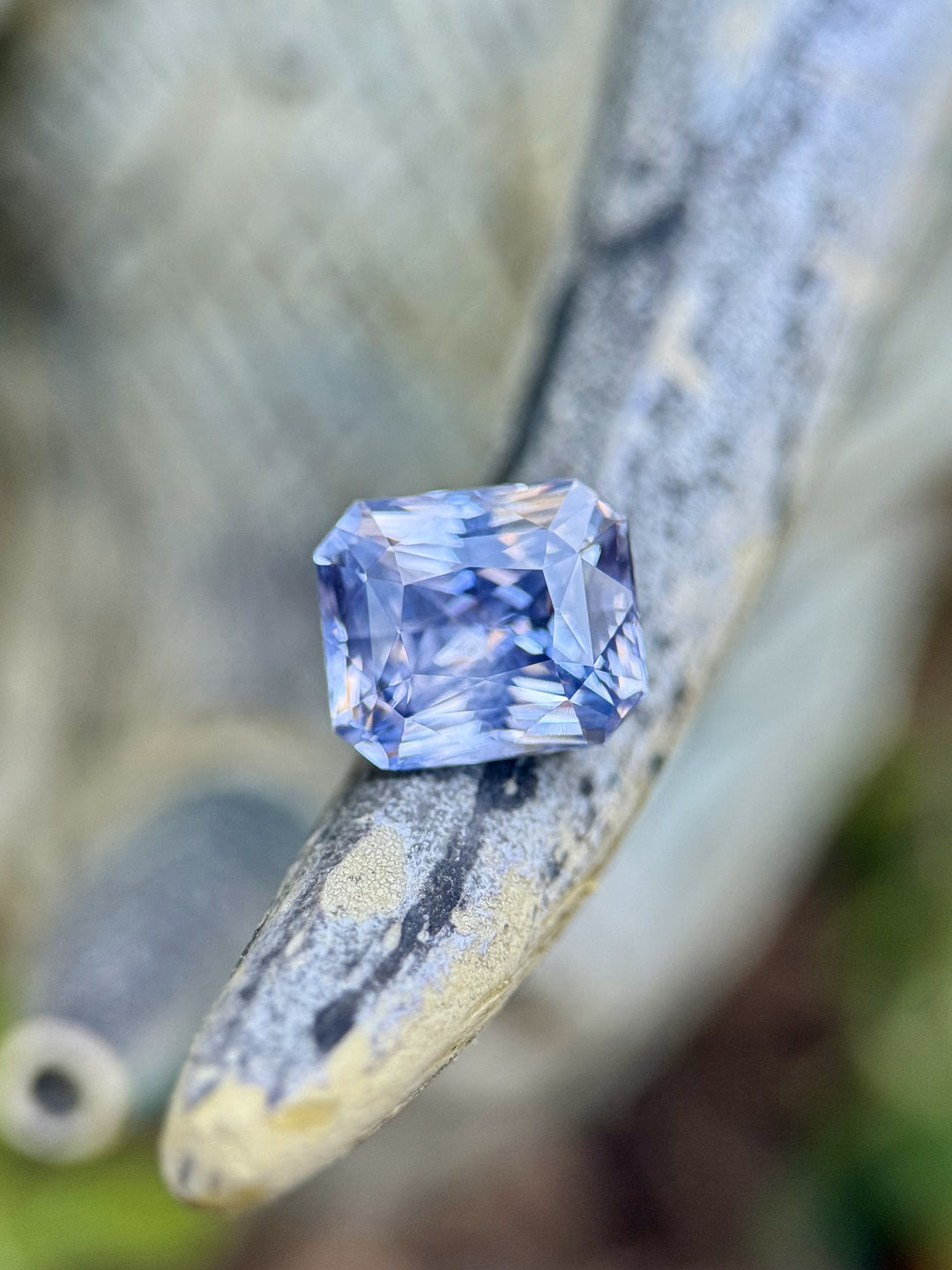 Sri Lankan 2.70-carat sapphire in radiant cut with soft violet and blue tones, resting on driftwood with a dreamy background.