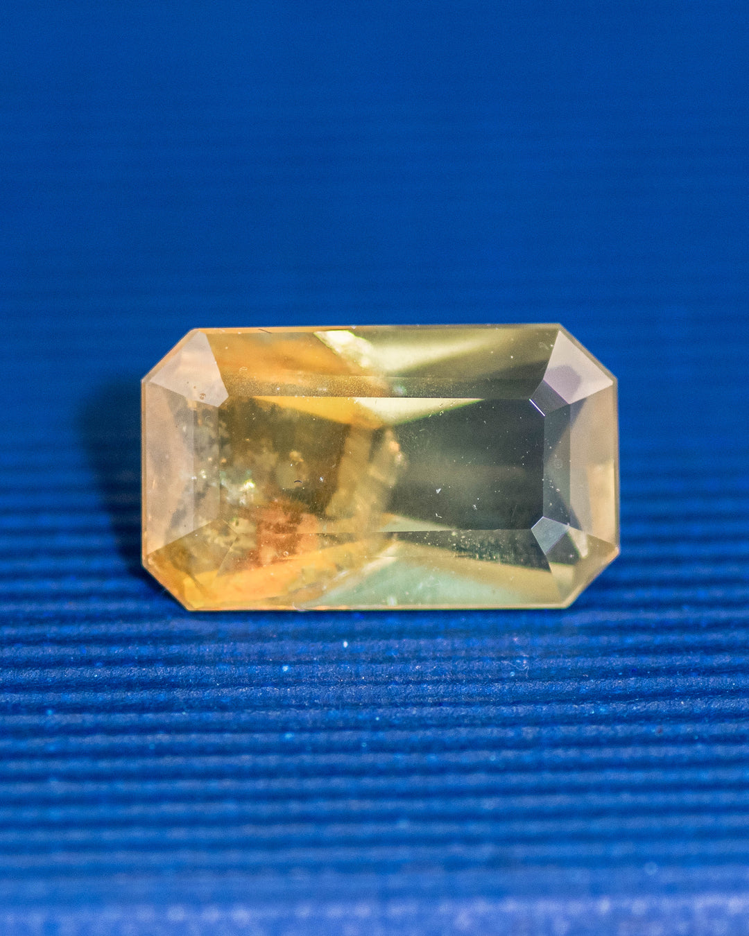 2.74 carat parti sapphire resting on a blue surface, displaying its natural mix of green, yellow, and orange hues with a custom octagonal step cut, untreated gemstone from Umba River, Tanzania.