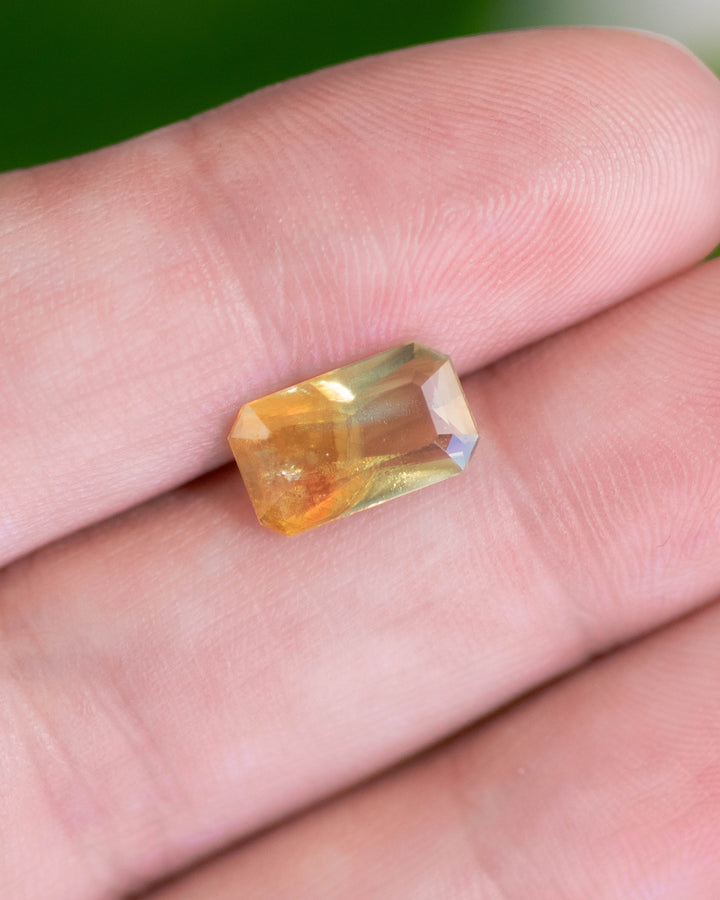 2.74 carat green, yellow, and orange parti sapphire displayed on fingertip under natural light, highlighting brilliance, clarity, and geometric symmetry of the custom octagonal step cut, untreated sapphire from Umba River, Tanzania.