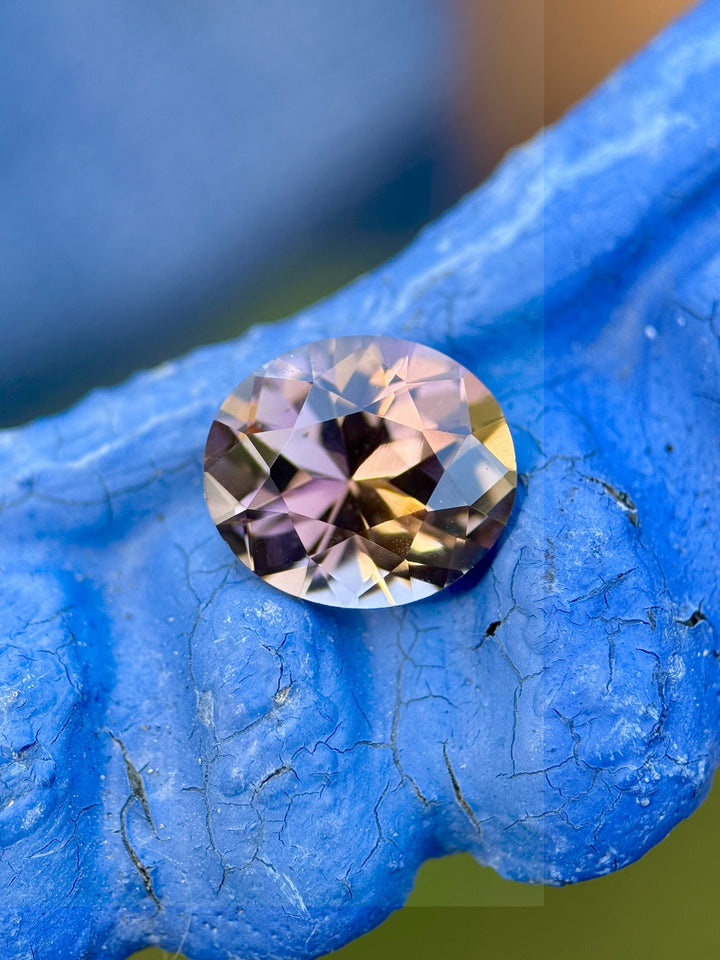 2.76-carat fancy Tanzanite gemstone displaying orangish peach, pink, and purple hues in a custom roval brilliant cut, photographed on textured blue background