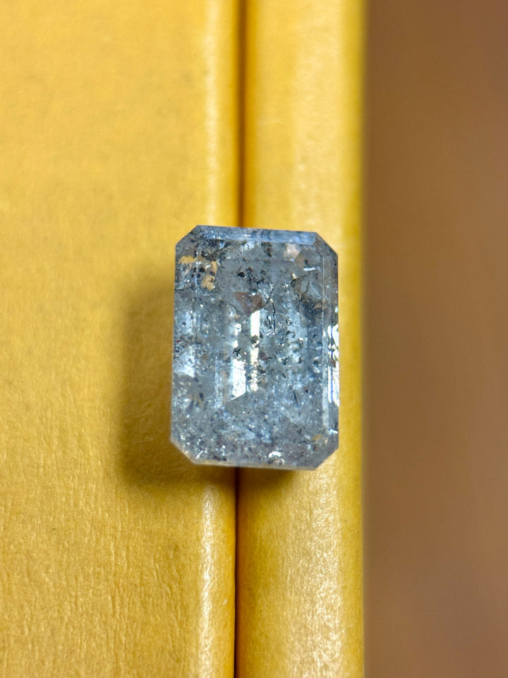 A rectangular emerald-cut blue-gray Diamond with visible internal inclusions, displayed against a textured mustard yellow background.