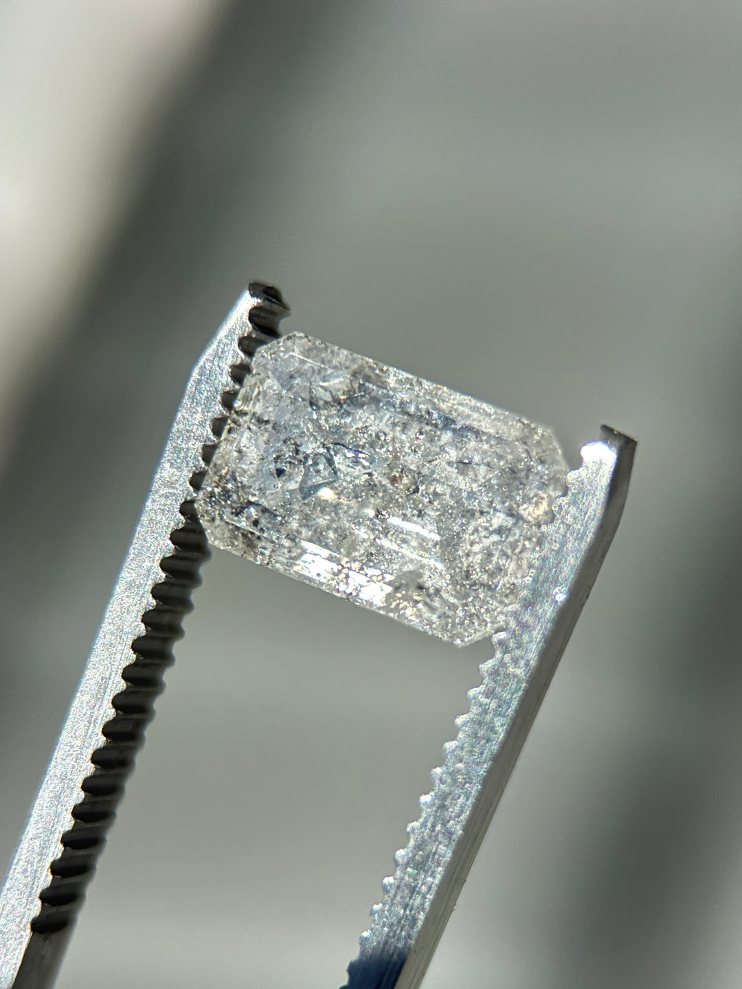 A close-up of a rectangular clear gemstone held in silver tweezers against a soft blurred gray background.