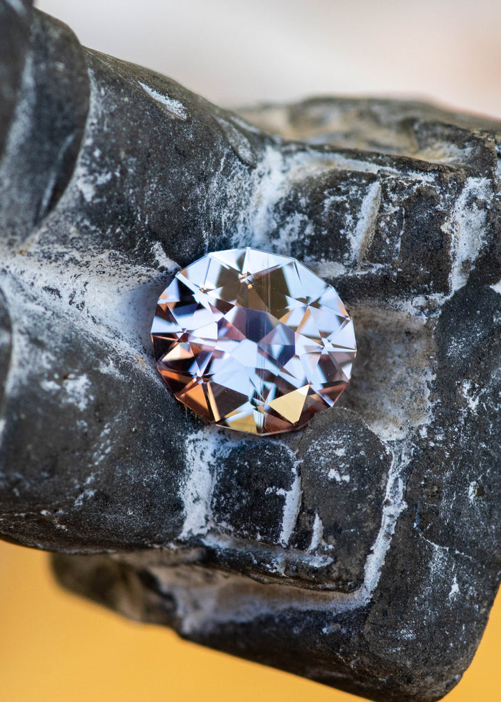 2.86ct peach to silvery violet color changing sapphire from Madagascar resting on stone carved figurine under natural light.