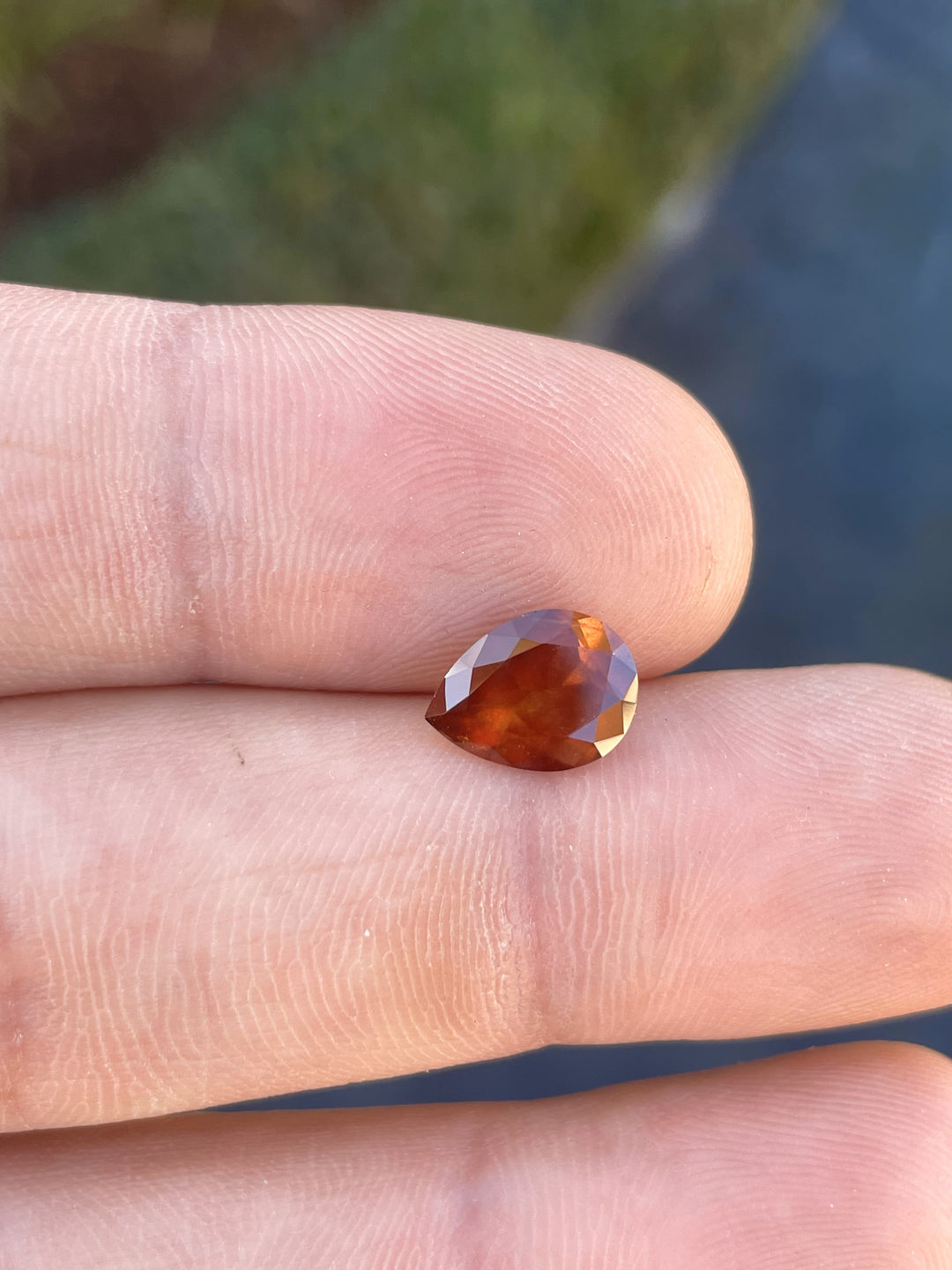 2.90 carat natural orange sapphire in a modified pear cut resting on a fingertip, highlighting its candy apple red and yellow hues.