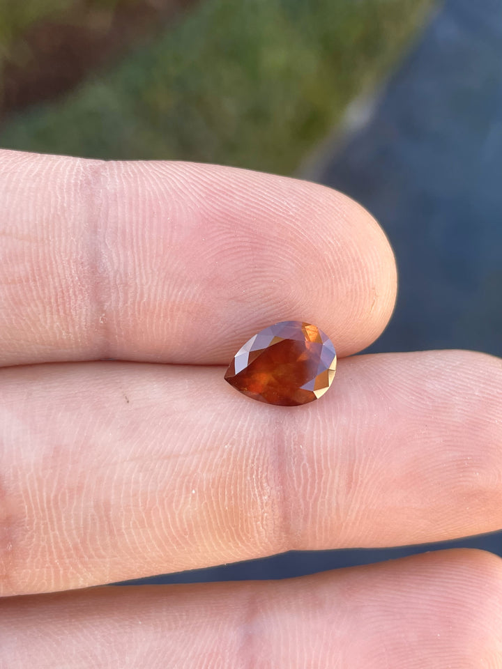 2.90 carat natural orange sapphire in a modified pear cut resting on a fingertip, highlighting its candy apple red and yellow hues.