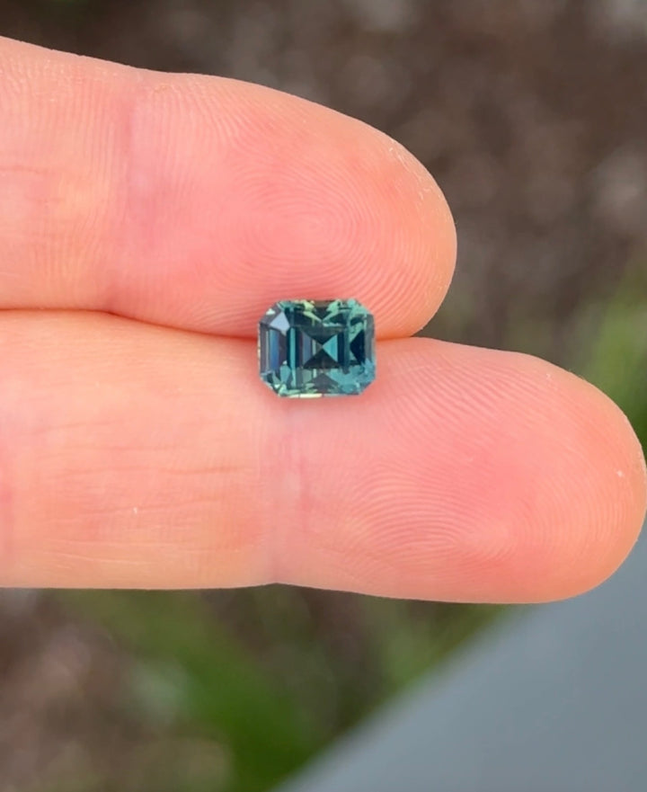 Green natural loose sapphire held between two fingers against a blurred natural background
