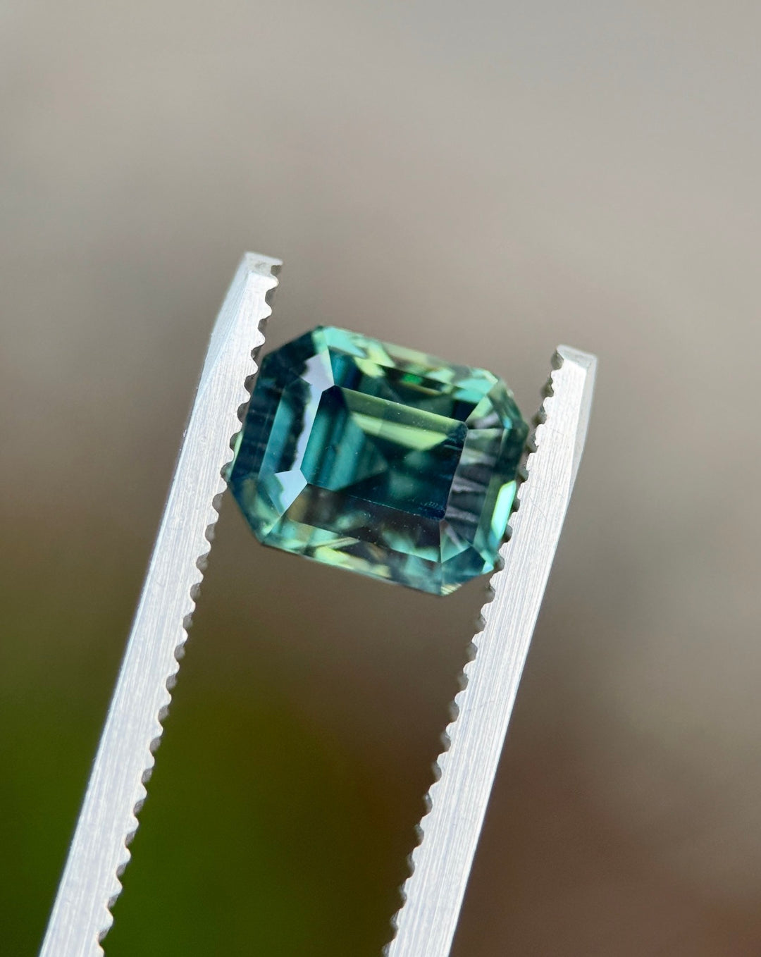 Loose teal sapphire held in gem tweezers highlighting natural green hues and precision faceting