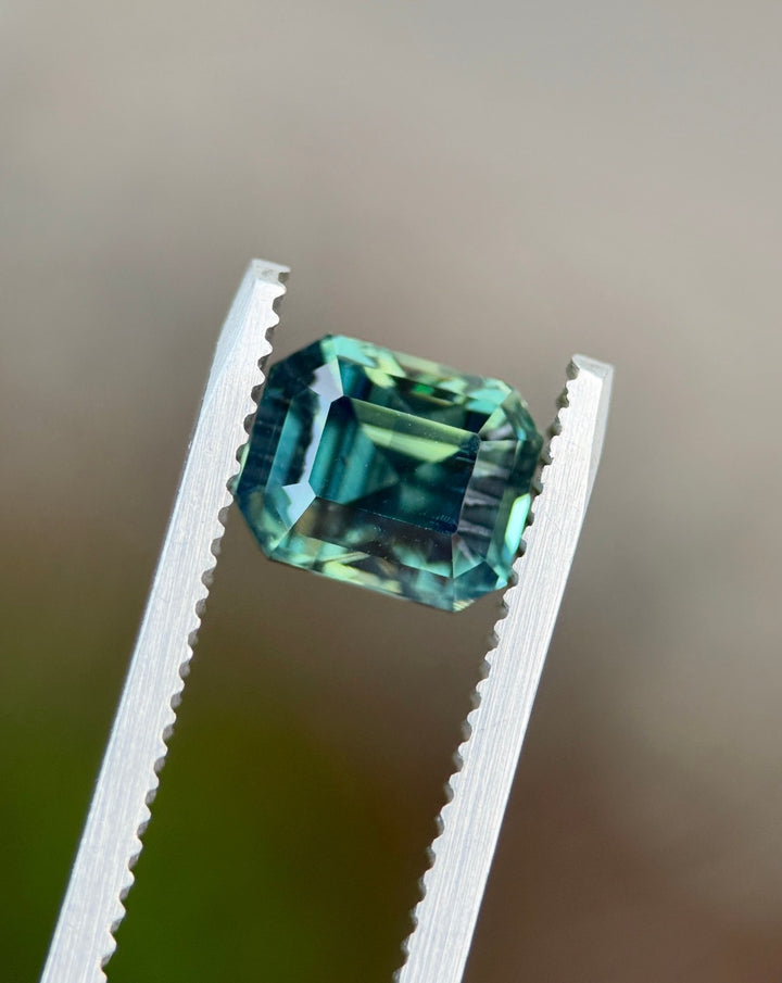Loose teal sapphire held in gem tweezers highlighting natural green hues and precision faceting