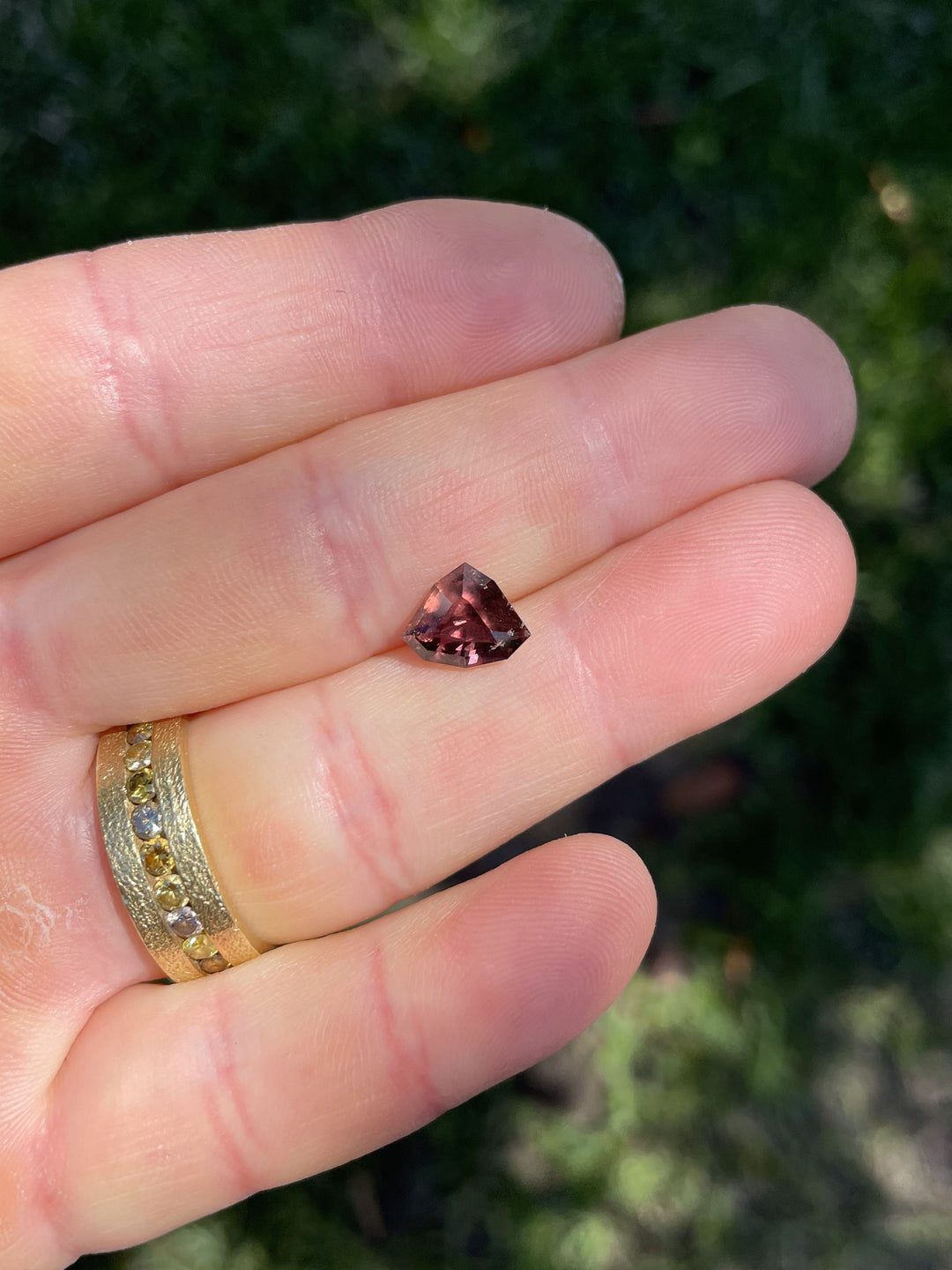 A 2.94-carat shield step-cut sapphire in rich strawberry red and dusty rose mauve tones, resting on a hand with a gold ring; natural light enhances the gem’s unique color and clarity.