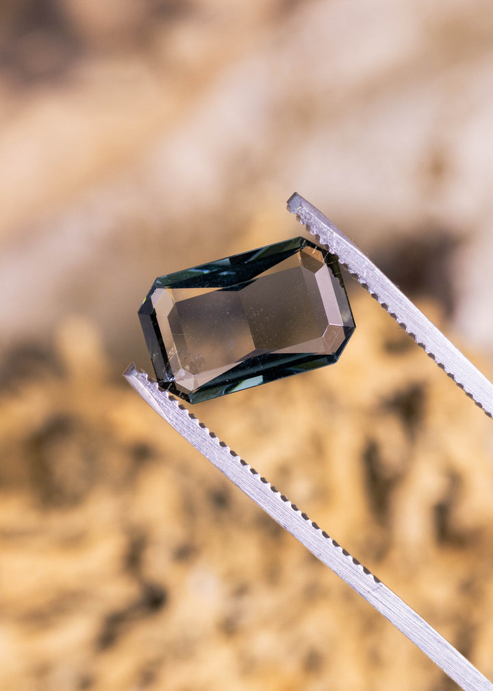 2.96 carat teal blue-green purple Tanzanite low profile emerald cut held in tweezers, precision faceted gemstone from Merelani Hills Arusha Tanzania displaying ultra-thin depth, crisp linear step facets and modern designer geometry