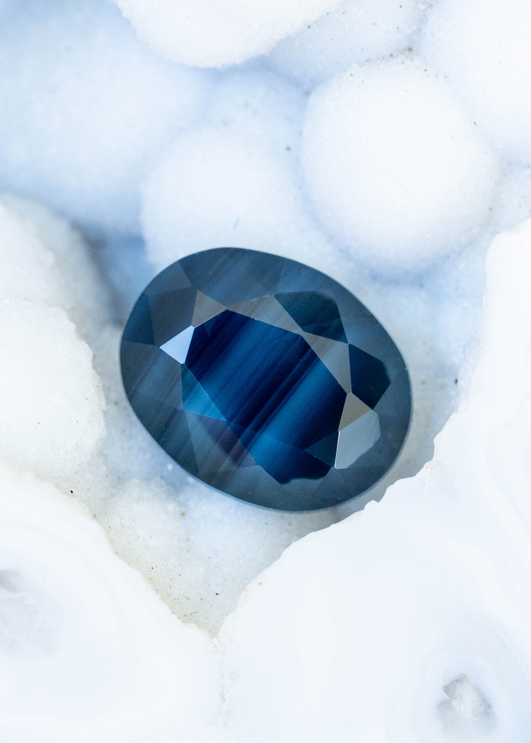 Oval sapphire resting on a textured white surface. A vivid cobalt-blue banded center is surrounded by smoky, deep blue facets, with fine parallel stripes visible across the gemstone.