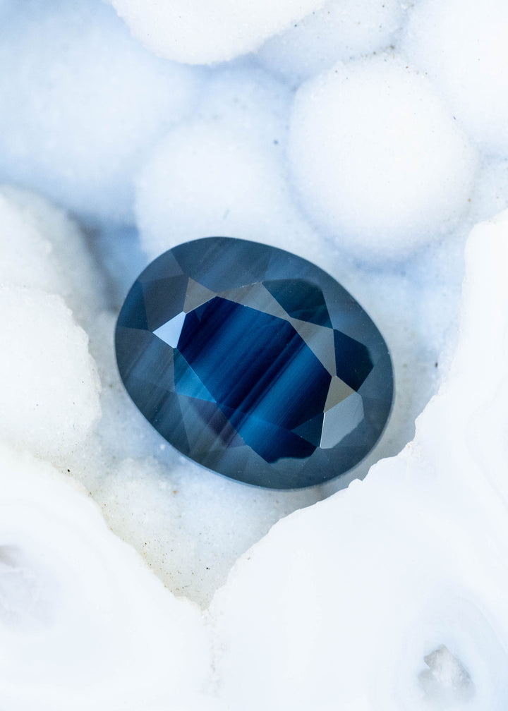 Oval sapphire resting on a textured white surface. A vivid cobalt-blue banded center is surrounded by smoky, deep blue facets, with fine parallel stripes visible across the gemstone.