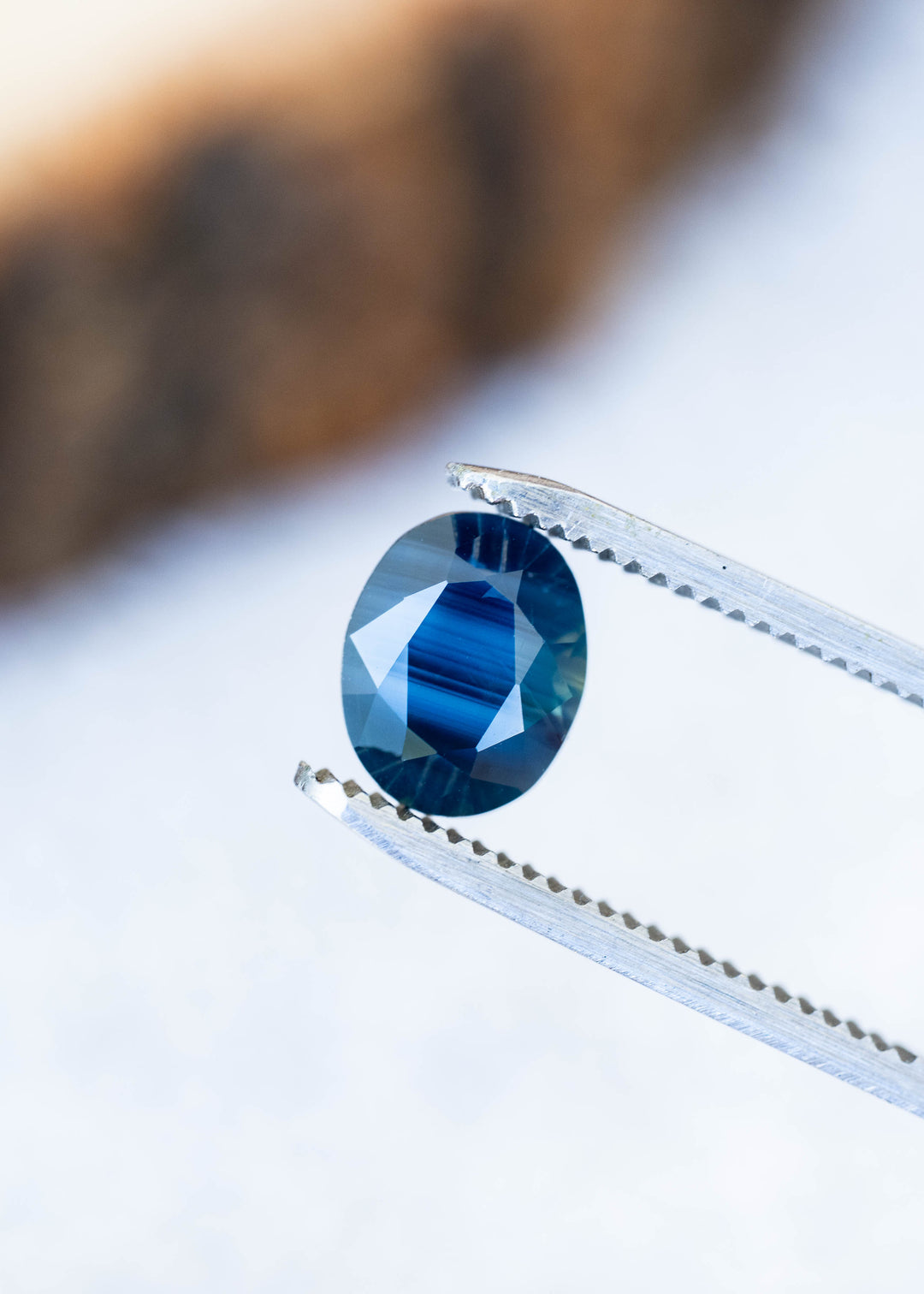 Deep blue oval sapphire held between silver tweezers against a softly blurred background. Bright blue parallel bands cross its center, bordered by darker blue facets and pale reflections.