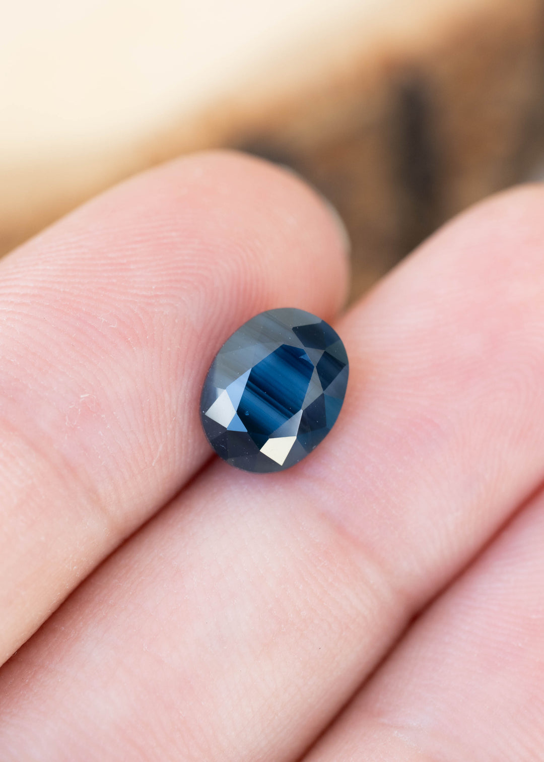 Oval sapphire resting between fingertips. Its center displays fine, vivid blue stripes, while the surrounding facets appear smoky navy blue with bright white reflections.