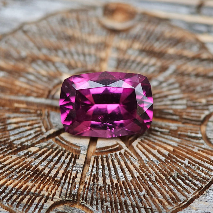 Stunning 4.29-carat garnet with a geometric modified cushion cut placed on an intricately carved wooden surface, exhibiting rich purple, pink, and red hues, sourced from Mombo, Tanzania.