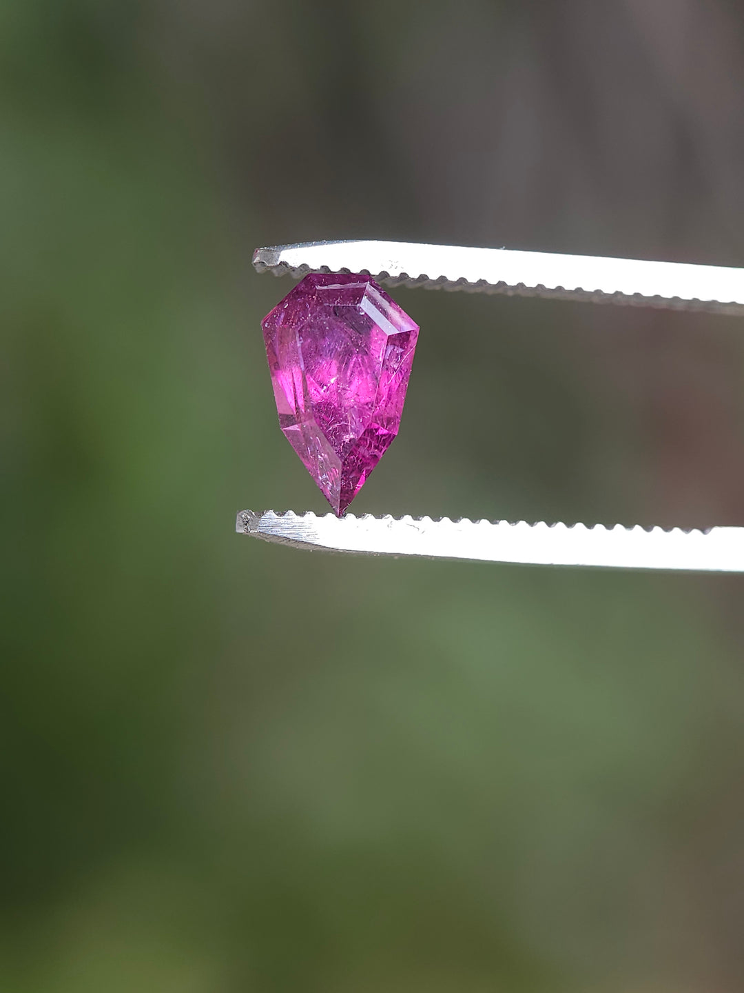 Close-up of a custom geometric shield-cut garnet held by tweezers, showcasing its vibrant blush to purple color, captured against a soft green blurred background.