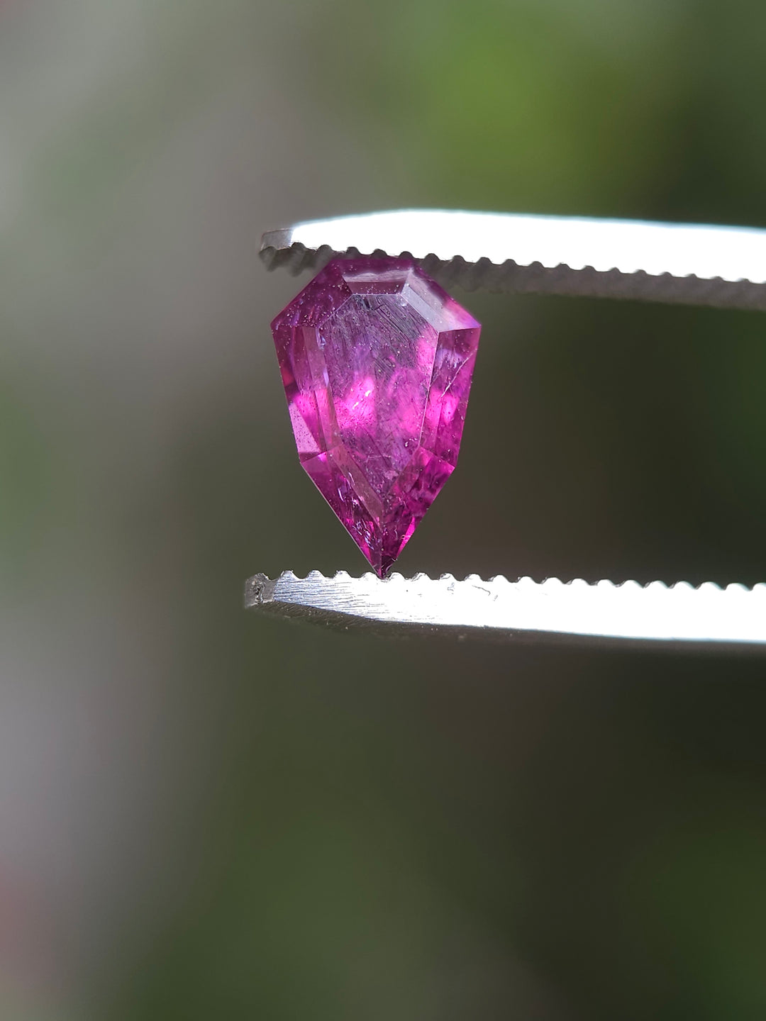 A 2.48-carat blush, purple, and red garnet gemstone in tweezers, illustrating the unique custom geometric shield shape and vivid color under natural light.