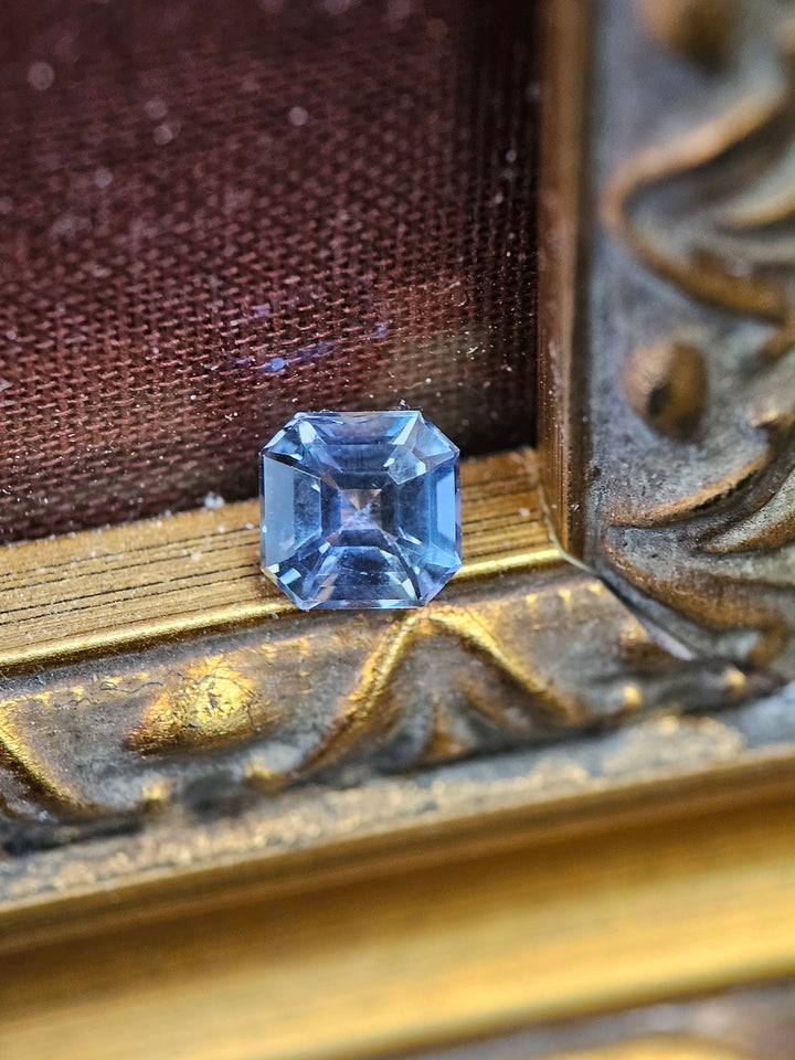 Teal, blue-green, and purple 1.10 carat Tanzanite gemstone in Asscher cut, beautifully displayed on a textured backdrop with intricate gold detailing.