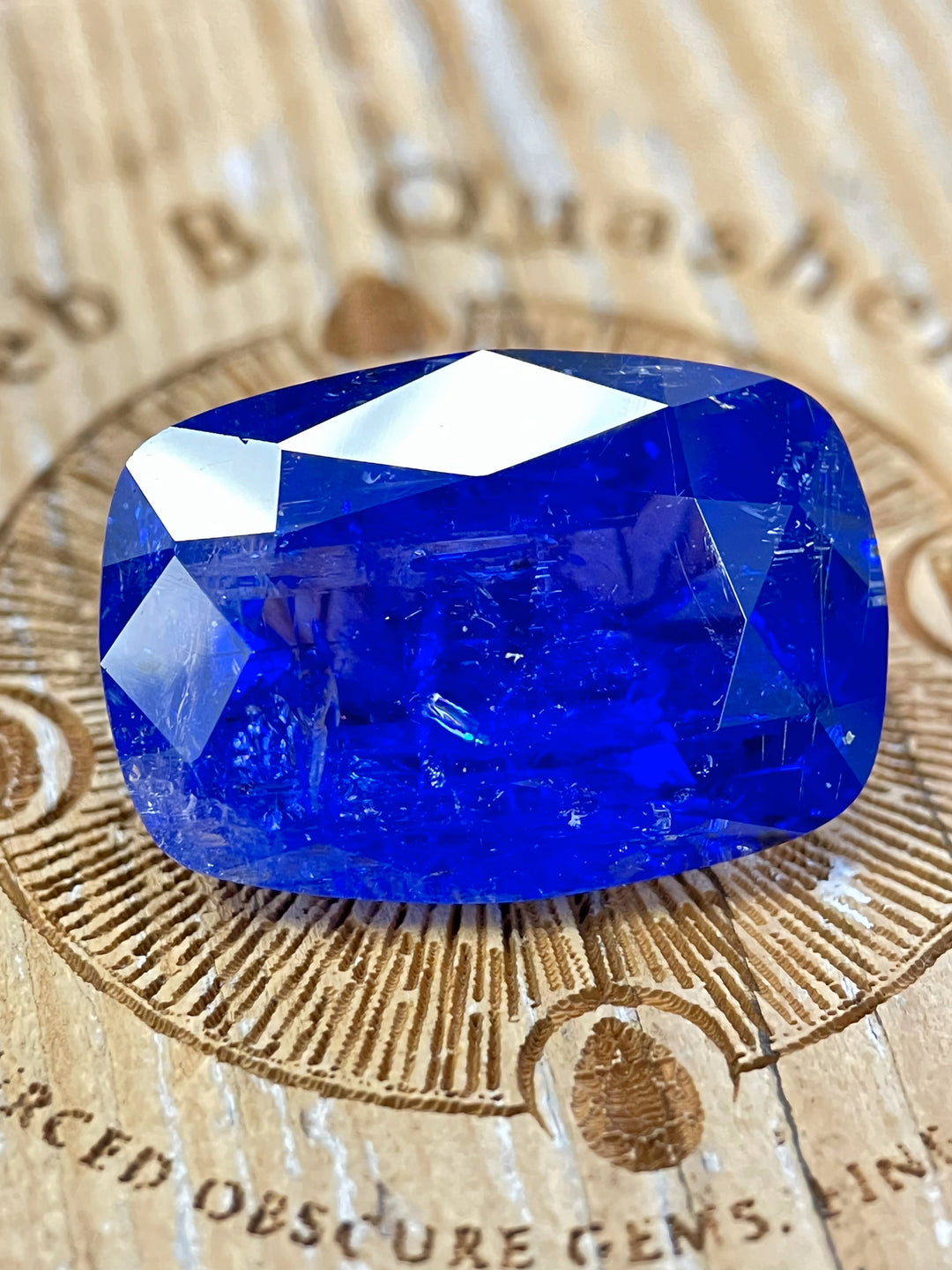 A stunning 27.31-carat cushion-cut tanzanite with vibrant royal blue and purple tones, resting on a wood background with engraved branding.


