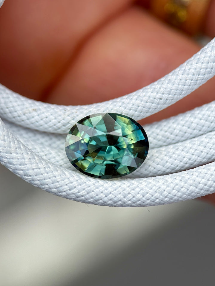 3.02CT Madagascar parti sapphire with yellow, teal, blue, and green hues displayed on white braided rope, highlighting the precision oval mixed cut and strong color shifts.