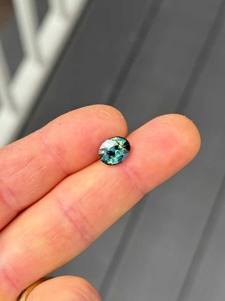 3.02 carat yellow teal blue green parti sapphire in a precision oval mixed cut resting on fingertips, showcasing vibrant color zoning and brilliance from Madagascar.