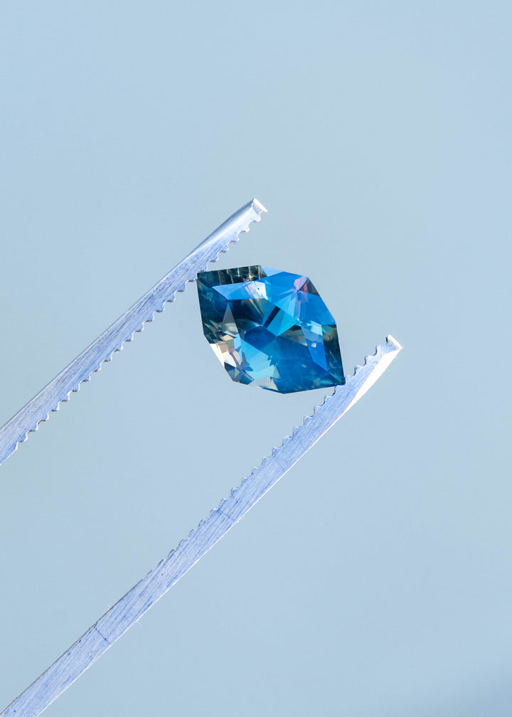 3.02ct opalescent teal parti marquise cut sapphire held in gem tweezers against gray background in natural lighting.
